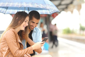 Interracial couple sharing a phone in a train station while wait under an umbrella in a rainy day